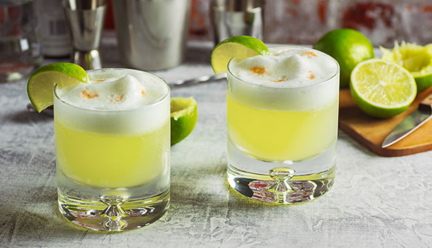 Two Pisco Sour Cocktails with Ingredients and Bottles on a Bar